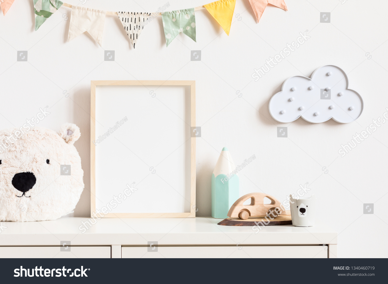 Stylish and modern scandinavian newborn baby interior with mock up photo or poster frame on the white shelf. Toys teddy bear wooden car and hanging cotton colorful flags and star. Template. Blank.