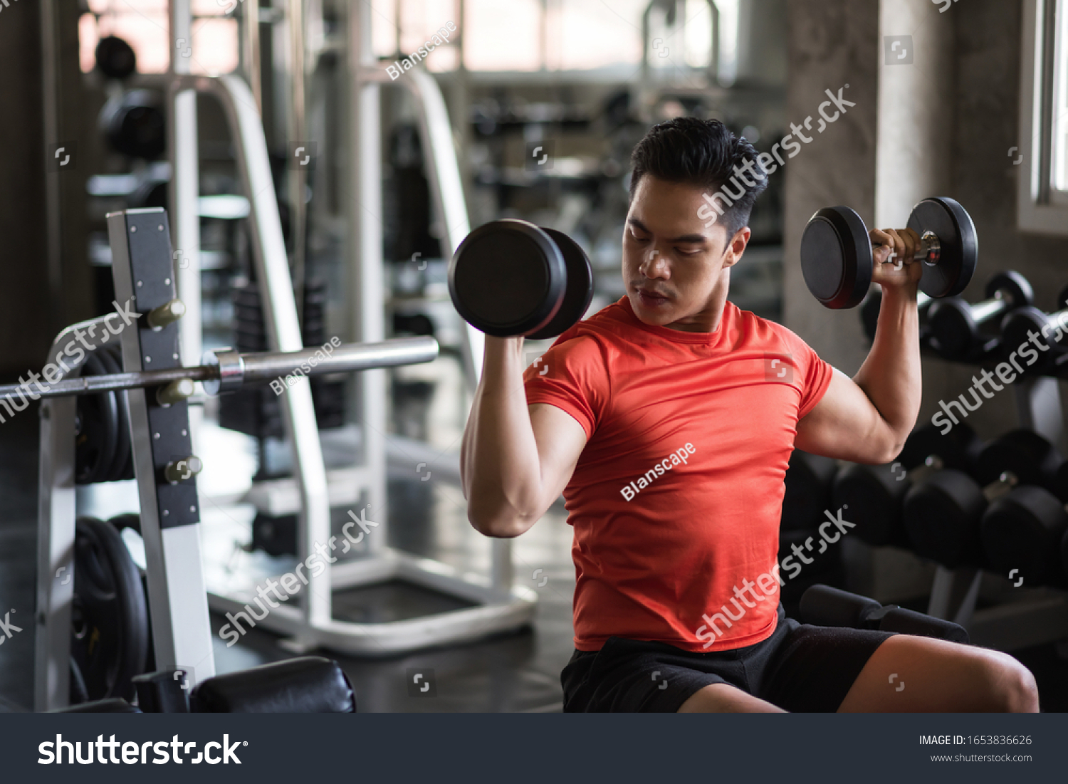 Strong Asian muscle man exercise biceps with two dumbbells with gym equipment background. Muscular body sportsman workout in sport fitness center. Bodybuilding and healthy lifestyle concept.