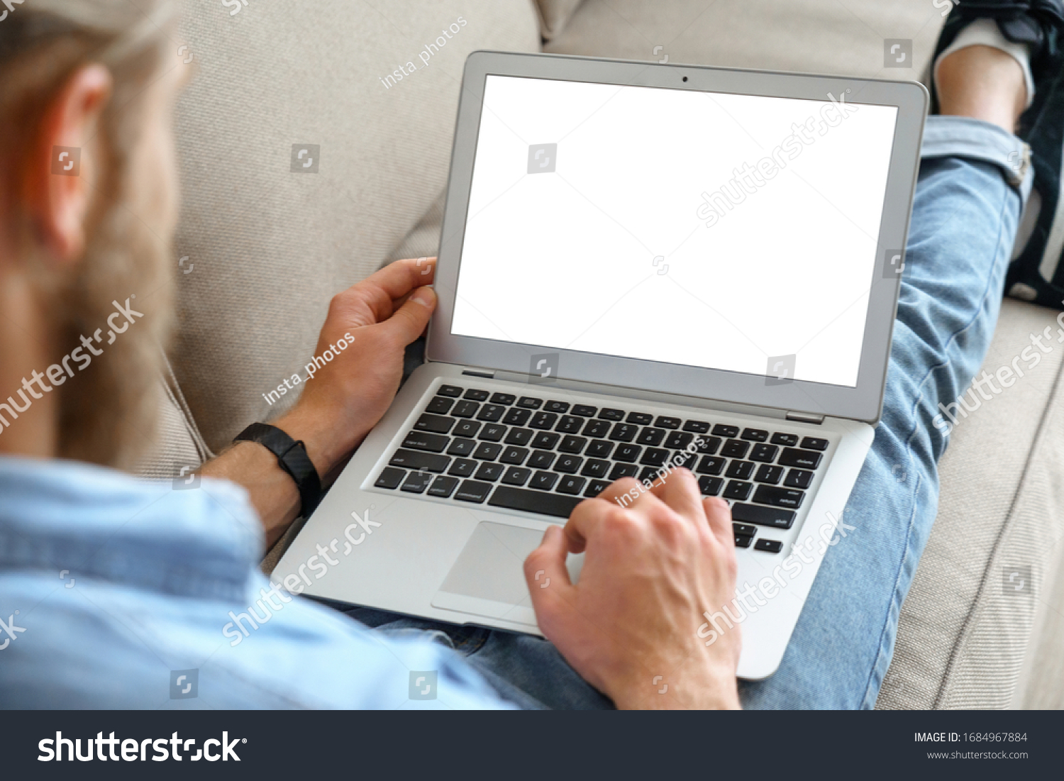 Young male tech user relaxing on sofa holding laptop computer mock up blank white screen. Man using modern notebook surfing internet read news distance online study work concept. Over shoulder view