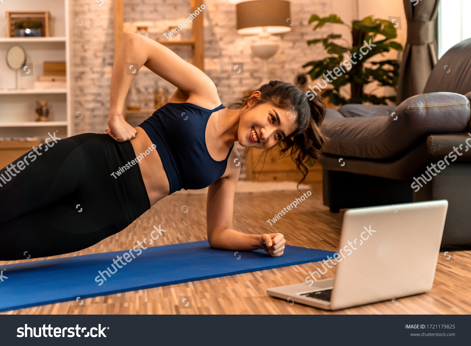 20s young Asian woman in sportswear doing stretching exercises while watching fitness training class on computer laptop online. Healthy girl exercising in living room with sofa couch in background.