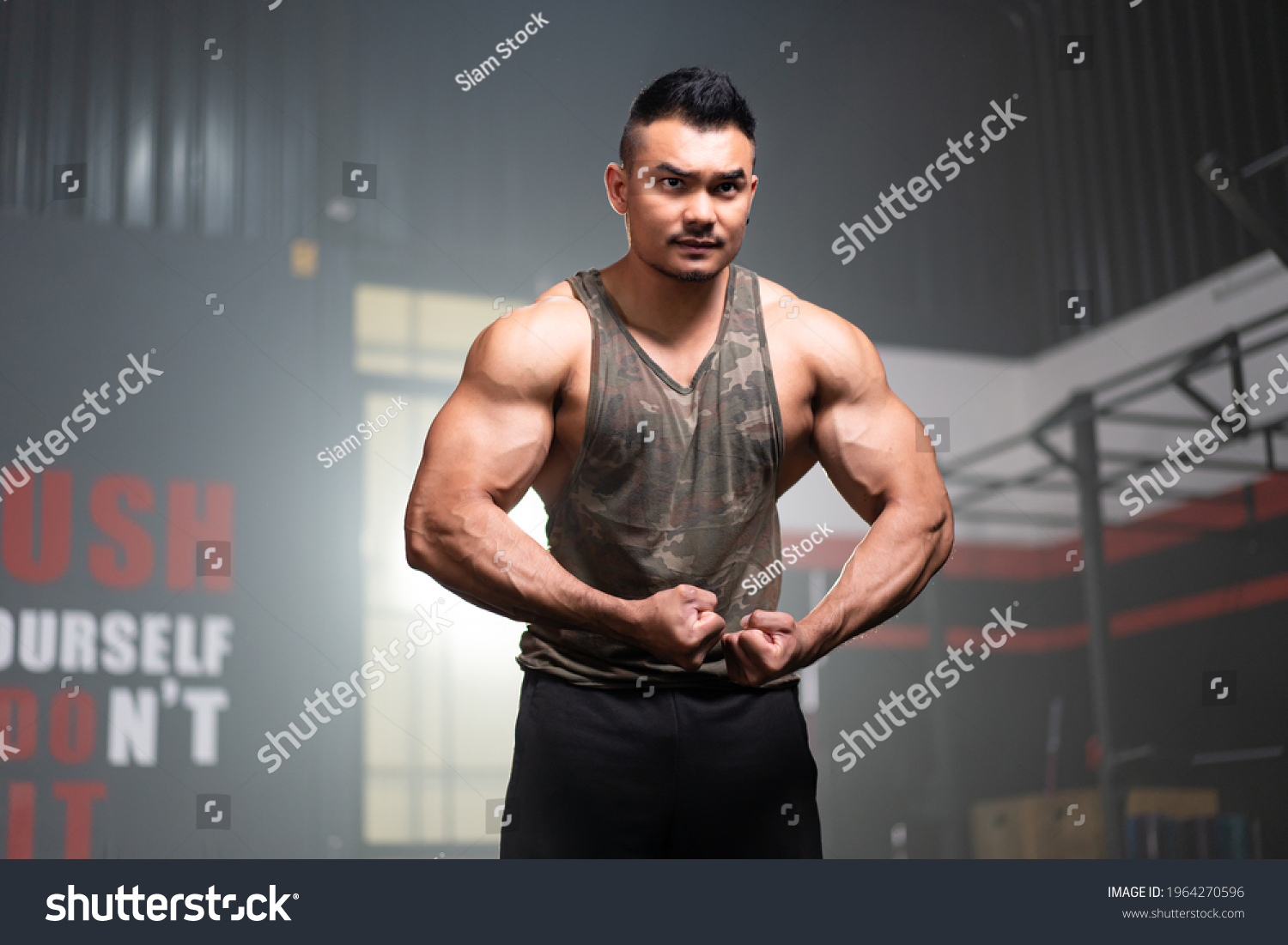 Young Asian muscular male fitness trainer in a tank top standing with hold his arms down flexing arm and shoulder muscles while looking forward with a serious face with a blurred background of gym.