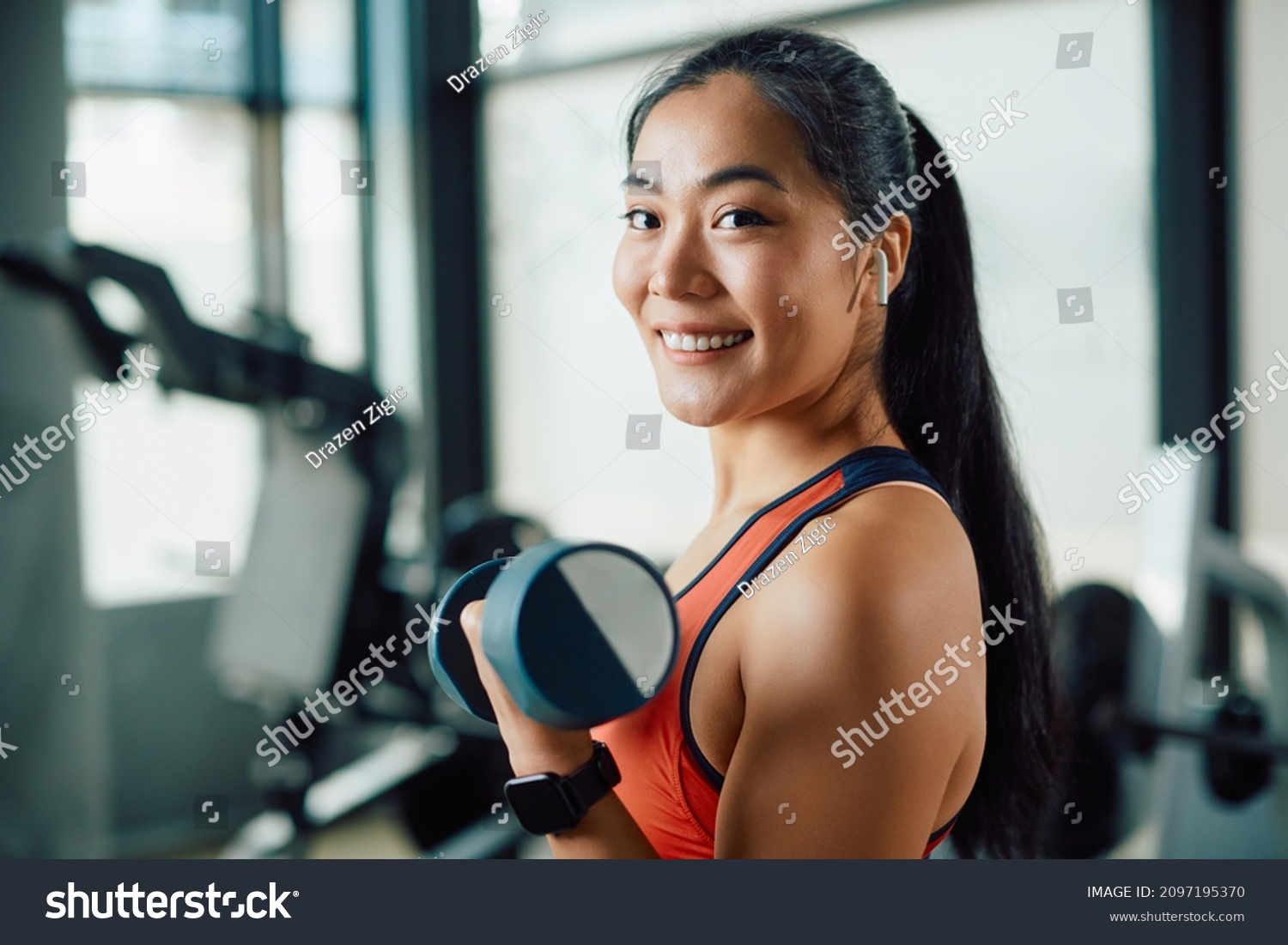 Happy Asian sportswoman lifting dumbbells during strength training in a gym and looking at camera.