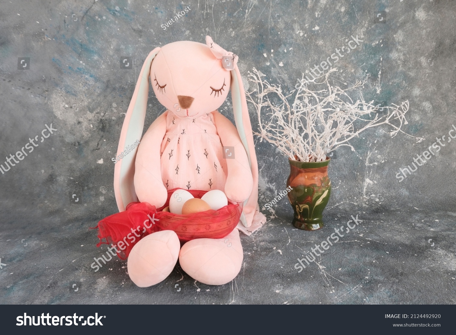 A pink sleeping hare a rabbit girl a toy with long ears sits holding a basket with white chicken eggs in her paws. Gray grunge background. Easter concept congratulations customs. Place for text