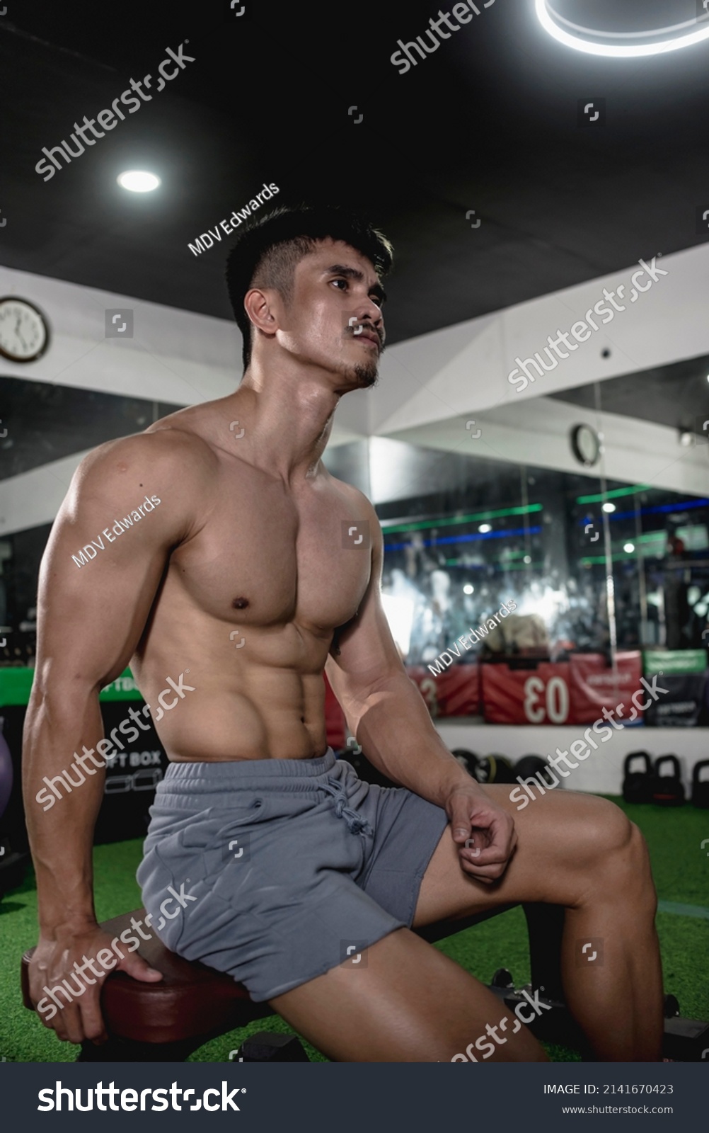 A shirtless asian man with a well built chest and a tight small waist sitting on a bench. A jacked male in his late 20s with an aesthetic physique at the gym. Wearing gray shorts.