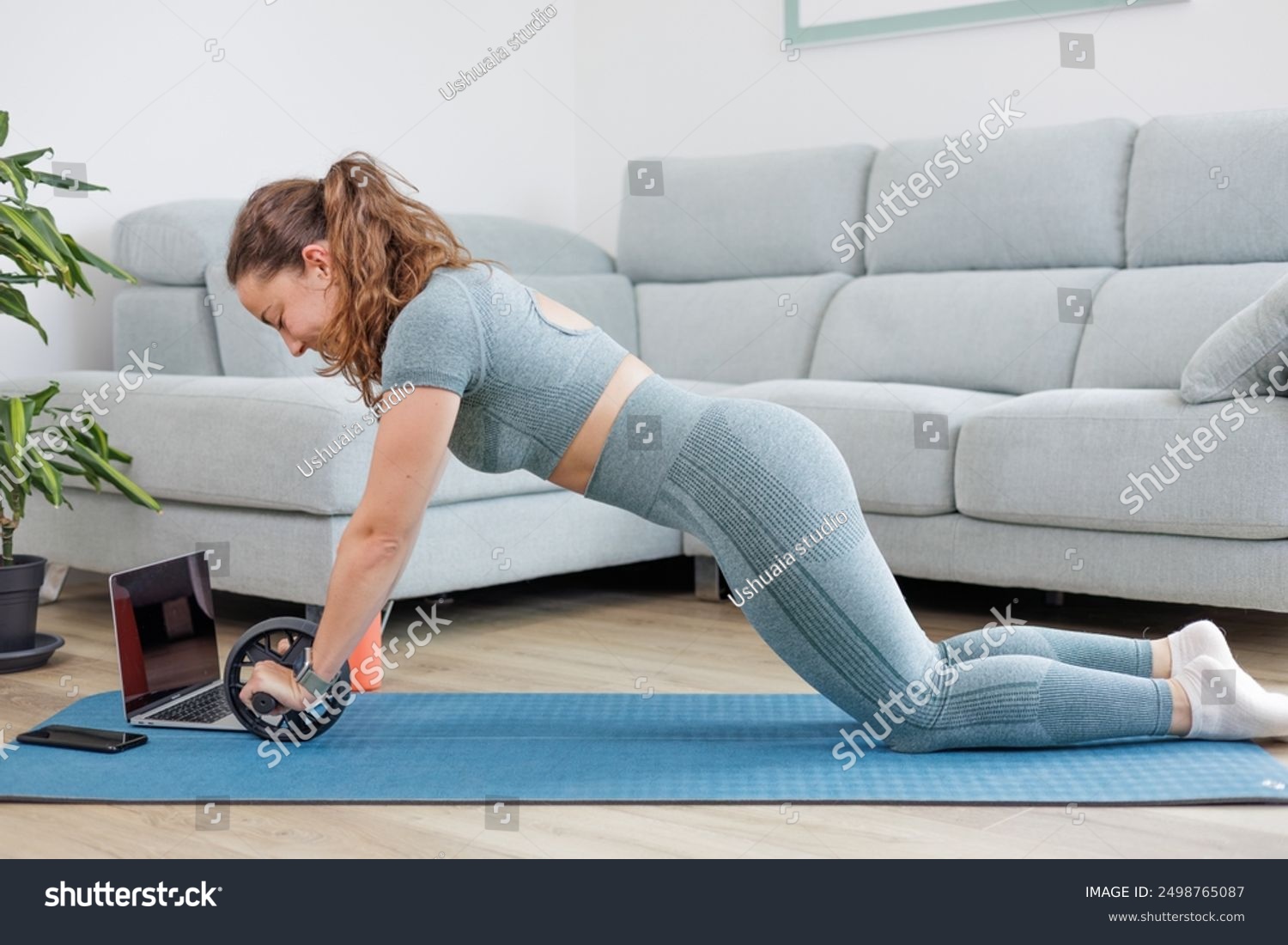 Young athletic woman does sports with a roller or wheel on a mat in the living room at home with an online personal trainer