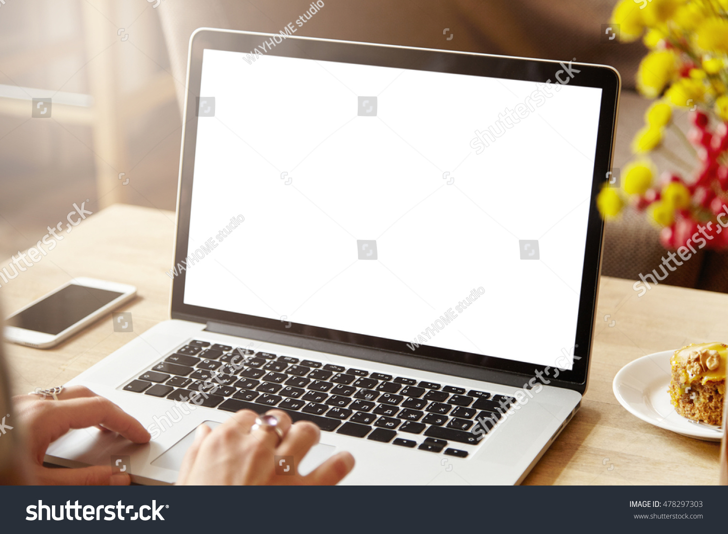 Woman's hands typing on laptop keyboard with white blank screen with copy space for your advertising content sitting at coffee shop table with mobile phone and cake next to her. Selective focus