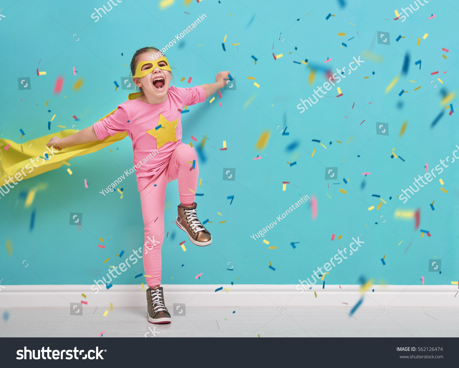 Little child plays superhero. Kid on the background of bright blue wall. Girl is throwing confetti and jumping. Yellow pink and turquoise colors.