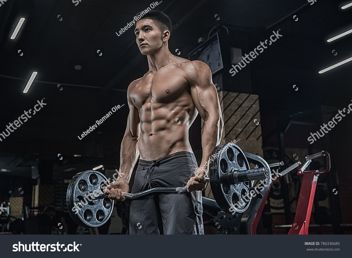 In a modern sports club an Asian man performs exercises for the biceps using a barbell against a dark background.