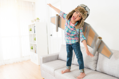 快乐开朗的女性的孩子在看相机笑，穿着古装的宇航员飞制作冒充站在沙发沙发上享受游戏时间。