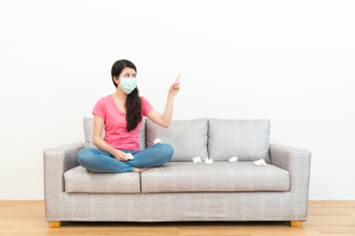 美丽的甜美女人戴着医用口罩，坐在在木地板沙发沙发上，指着白色背景想着生病的解决方案。