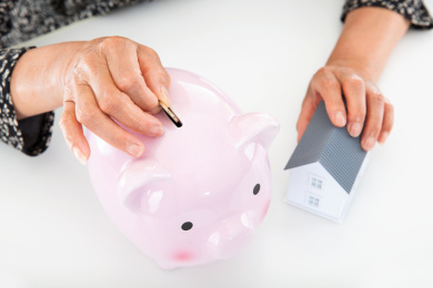 一位老太太一只手拿小房子模型另一只手把一枚硬币放入小猪存钱罐