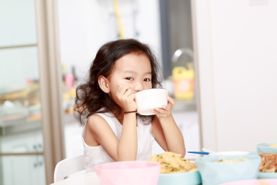 在家里餐桌前端着小碗吃饭的小女孩