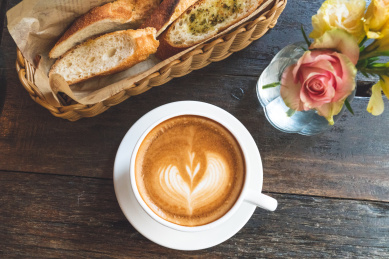 餐桌上放着一杯爱心形拉花的咖啡、蒜香法棍、鲜花
