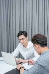 在白色办公桌及银色笔记本电脑前讨论工作的两位年轻男性白领