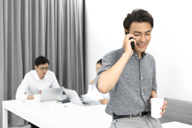 一名商务男士靠在办公桌上接打电话后面的两名同事认真工作