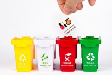 白色背景上放着一排彩色垃圾桶，一只手拿着代表不同种类垃圾的卡片往垃圾桶里面丢，用玩具组合来表达垃圾分类的概念