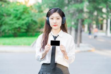 美女大学生拿着手机在学校内指着手机带着戴着耳机听听力