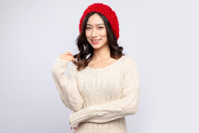 灰色背景里，身穿米色毛衣的带着红色绒线帽的美丽亚洲女孩