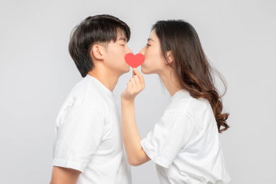 灰色背景里，手里拿着爱心纸片正在亲吻的亚洲年轻情侣