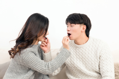 冬天一对高兴的亚洲年轻情侣坐在沙发上，互相嘴对嘴，亲密的吃着巧克力